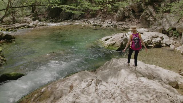 Young tourist girl wearing straw hat, with pink backpack walking to mountain river