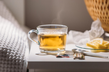 Influenza, flu season, cold concept. Close up view of cup of hot green tea, pills, digital thermometer and saucer with lemon slices on bedside table, tissues and nightlight on background. 