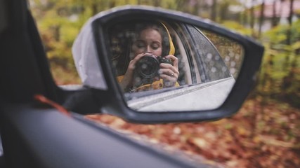 Woman Making a Self Portrait Video in a Rear View Mirror in a Driving Car. SLOW MOTION. Young artistic girl making a video with digital camera, travelling in a car through rainy autumn forest.