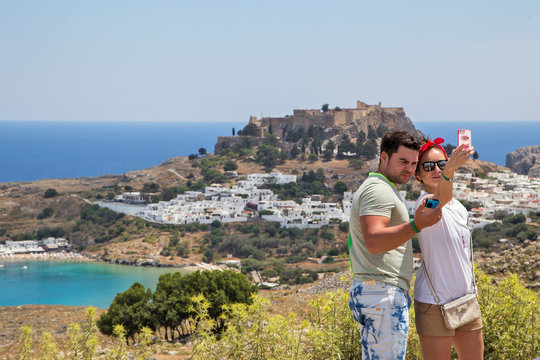  A man and a woman stand on a hill and take a selfie against the city of Lindos .Horizontally.