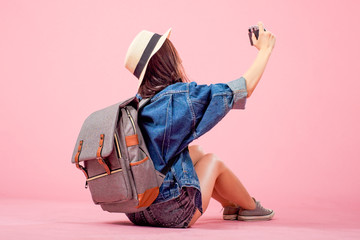 Smiling woman traveler sitting holding camera with backpack in holiday on pink backgrounds,...