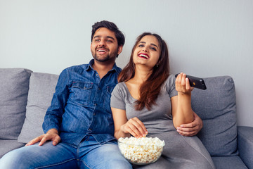 young indian couple watching comedy movie with remote control in hand in living room with popcorn