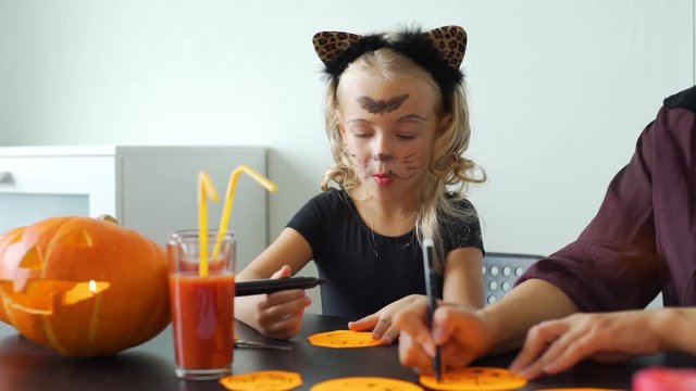 Little Girl with Mom Preparing for Halloween Celebration. They are Drawing Scary Faces on a Colored Paper. Holidays and Halloween Decorations Concept