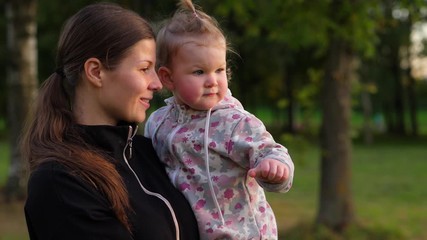 Woman with little baby on hands, look together to someone and wave hand in farewell. Slow motion portrait shot of mother with small kid. Green outdoors of park, evening hour