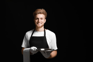 Handsome waiter with empty tray on dark background