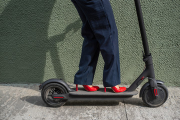 Obraz premium Young woman in formal wear on red hight heels is standing on electrical scooter. Close-up of female legs. A business woman in a trouser suit and red shoes moves around the city on an electric scooter.