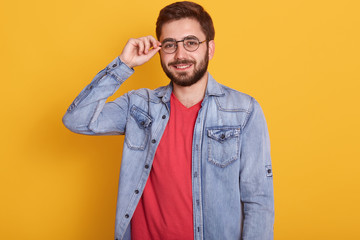 Studio shot of young attractive handsome man wearing red t shirt and denim jacket standing against yellow wall and smiling, keeps hand on his spectacles, looking directly at camera, bearded male.