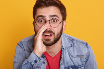 Horizontal shot of handsome man wearing casual red t shirt and stylish demin jacket, keeps hand on mouth telling secret rumor, whispering malicious talk conversation, looking directly at camera.