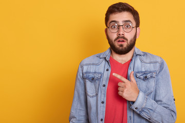 Image of shocked young man pointing finger aside at blank copy space, astonished surprised guy dressescasual clothing, looking directly at camera, standing isolated over yellow studio background.