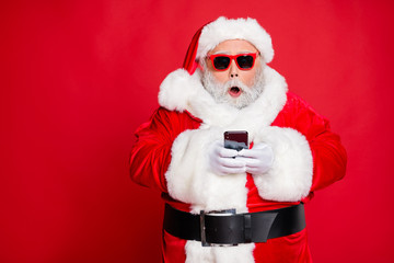 Portrait of impressed christmas father in hat using cell phone device screaming omg wearing read about festive events eyeglasses eyewear belt isolated over red background