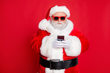 Fototapeta na wymiar Portrait of minded santa claus with eyewear eyeglasses using cell phone device reading news wearing costume isolated over red background