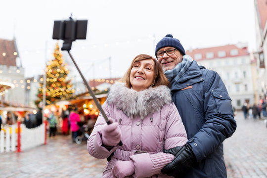 technology, winter holidays and people concept - happy senior couple taking picture by smartphone on selfie stick at christmas market on town hall square in tallinn, estonia