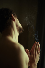Handsome bearded male with muscles practicing yoga  indoors, using incense stick for calm and...