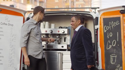 Medium tracking shot of middle-aged Arab businessman walking down city street towards mobile coffee van, greeting young Caucasian barista, making order and chatting while beverage is being prepared