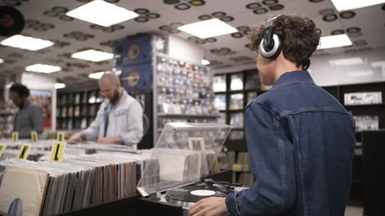 Waist-up shot of Caucasian female music fan standing by turntable in record shop and playing vinyl disc, listening through headphones and dancing enthusiastically, and customers browsing in background