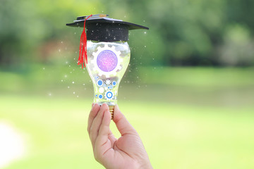 Woman hand holding cog inside in light bulb with graduates hat on natural green background,...