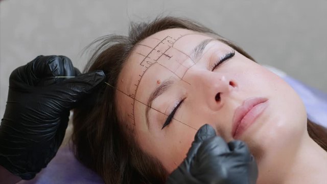 A face of a young lady and hands of a microblading master in black gloves. A specialist is marking the highest top of an eyebrow with a thread, a client is lying with closed eyes. Close up, portrait.
