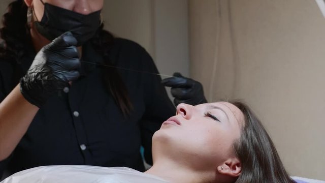 A microblading master in mask is measuring young woman's eyebrows in the beauty studio. A lady is lying with closed eyes and a specialist is making marks with a thread for a perfect shape. Close up.
