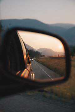A rearview mirror reflecting a landscape with mountains, a sunset and a road. 