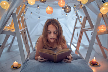 Little 10 year old girl reading classic book under her home-made tent inside the living room.
