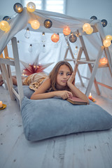 Little 10 year old girl reading classic book under her home-made tent inside the living room.