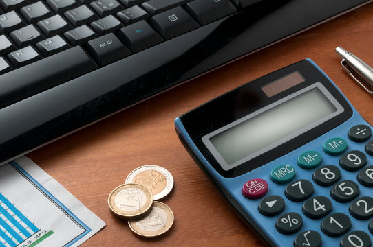A calculator on wooden table with a pen and some euro coins in front of a black computer keyboard