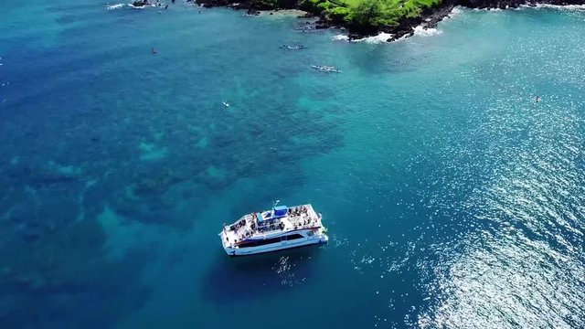 A drone, birds eye, aerial view of a party boat on the the beautiful teal waters along the coast of Wailea beaches on the island of Maui, Hawaii. Paddle boats, beautiful homes & resorts come into view