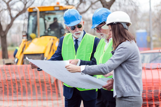 Mixed team of senior men and young female architects or business partners looking at building plans on a construction site. Engineers meeting and discussing blueprints while visiting work site
