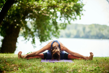 Naklejka na ściany i meble Dedicated Caucasian woman sitting on mat in nature near water and doing Seated Wide Legged Forward Bend yoga pose.