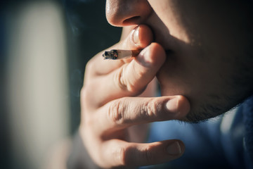 A young man in blue clothes and with a stubble on his face takes a drag from a Smoking cigarette, which he holds with his fingers.