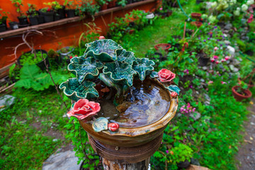 A beautiful small decorative fountain made of porcelain or clay molded in the form of green leaves and rose flowers through which water flows for drinking in a garden with green plants.