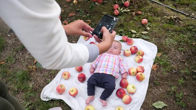Mother takes mobile phone picture of infant baby daughter lie in apples on rug in fruit garden for social media