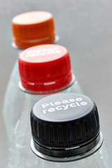 Plastic bottles and black, red and orange bottle caps with please recycle message against a grey background. Concept of plastic pollution. Vertical image.