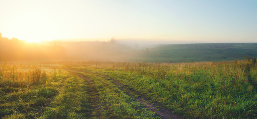 Sunny summer landscape with country road and green hills covered by morning fog