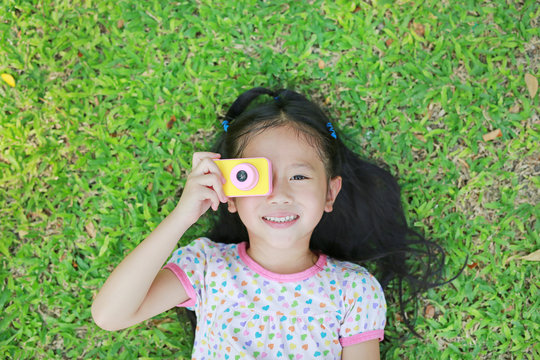 Cheerful little Asian girl takes photo with colorful digital camera lying on green lawn background.