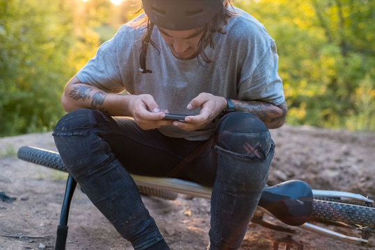 young cyclist guy , with tattoos, sitting on a bicycle in a protective helmet, uses the Internet in a smartphone, on a warm natural background. active lifestyle