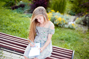 Naklejka premium beautiful little girl child, sitting on a bench in a summer park, with flowing hair, in a dress, leafing through a notebook. School lifestyle