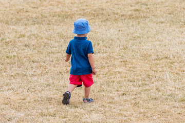 Naklejka na ściany i meble Lonely little boy walking on dry grass of field or stadium_