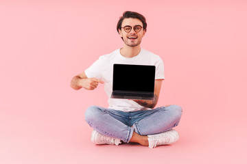 Image of happy handsome man wearing eyeglasses winking and pointing finger at laptop while sitting on floor