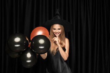 Young blonde woman in black hat and costume on black background. Attractive, sensual female model. Halloween, black friday, cyber monday, sales, autumn. Copyspace. Holding balloons, whispers secret..