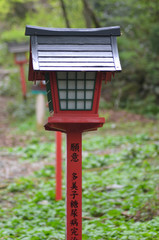 A black lantern with a red pole is in a park. Japanese lettering in black is on the pole. Other lamps are in the distance, with trees in the background.