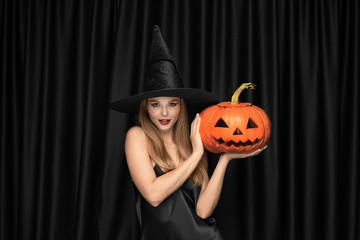 Young blonde woman in black hat and costume on black background. Attractive, sensual female model. Halloween, black friday, cyber monday, sales, autumn. Copyspace. Looks mystical, holding pumpkin.