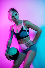 Fit young blonde woman posing with medicine ball in gym in neon lights.
