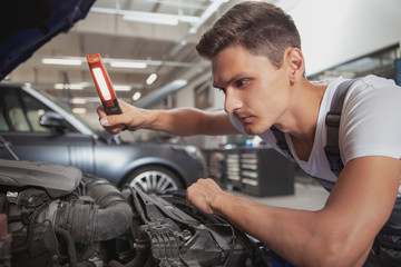 Close up of a concentrated young male mechanic looking under the hood of an auto at the workshop. Handsome car service worker repairing broken automobile