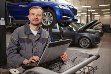 Cheerful mature male car mechanic smiling to the camera, working on a computer at his garage. Happy auto technician repairing automobile at his workshop