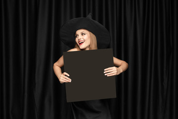 Young blonde woman in black hat and costume on black background. Attractive, sensual female model. Halloween, black friday, cyber monday, sales, autumn. Holding black sheet for copyspace, looks mystic