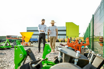Young agronomist walking with salesman at the open ground of the shop with agricultural machinery, choosing new plow for tillage