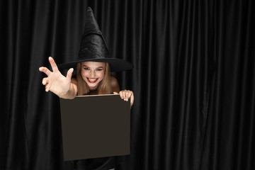 Young blonde woman in black hat and costume on black background. Attractive, sensual female model. Halloween, black friday, cyber monday, sales, autumn. Holding black sheet for copyspace, looks mystic