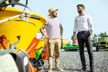 Young agronomist with salesman at the open ground of the shop with agricultural machinery, buying a new planter for farming