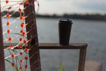 A paper cup of coffee left on a wooden handrail at the railing by the lake